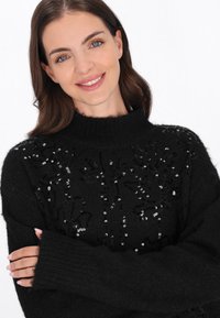 Czarny sweter z wysokim kołnierzem i długimi rękawami, z teksturowanym splotem i cekinowymi wzorami kwiatowymi na przodzie. Miły, przytulny wygląd.