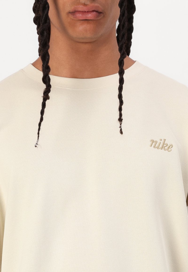 T-shirt en tricot crème avec un col rond et des manches courtes ; présente un petit logo doré "Nike" brodé sur la poitrine gauche.