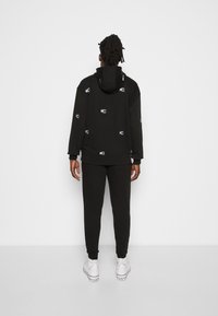 Felpa nera con cappuccio e logo bianco discreto, abbinata a pantaloni jogging neri e sneakers bianche, evidenziando uno stile casual e atletico.