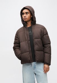 Καφέ κοντό φουσκωτό μπουφάν με εμπρός φερμουάρ, ευρύχωρη κουκούλα και τμηματική ραφή κατά τετράγωνα. Συνδυασμένο με μαύρο υφασμάτινο πουκάμισο και ανοιχτό μπλε τζιν.