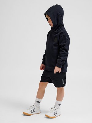 Barn stående i siden iført en sort hættetrøje, sorte shorts, hvide sokker og hvide sneakers med gummisåler.