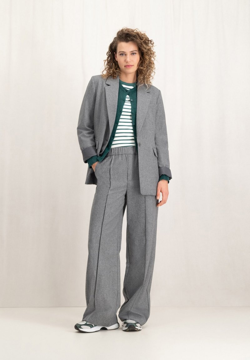 Grijs wollen mix pak met wijduitlopende broek en een getailleerde blazer, gelaagd over een groen gestreept sweater. Sneakers maken de look af.