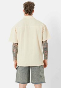 Kurzärmliges, cremefarbenes Hemd aus strukturiertem Stoff mit lässiger Passform. Es hat einen klassischen Kragen und einen geraden Saum. Getragen mit grauen Denim-Shorts.