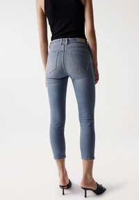 Jeans de denim ajustados en azul claro con cintura alta, longitud recortada y dos bolsillos traseros. Combinados con sandalias negras de tacón.