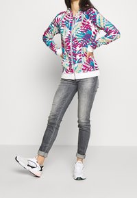 Colorful zip-up hoodie with a floral pattern in pink, blue, and green. Paired with light gray jeans and white sneakers.