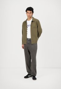 Olive green button-up jacket with chest pocket, white graphic t-shirt, gray wide-leg pants, and black sneakers with three white stripes.