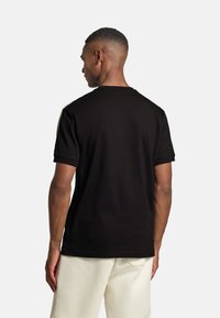 Man med kort hår som bär en svart kortärmad skjorta och vita byxor, sedd bakifrån mot en enfärgad vit bakgrund.