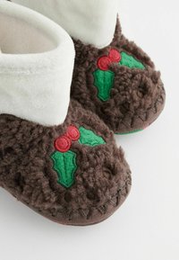 Braune, flauschige Hausschuhe mit weißen Bündchen, die rot gestickte Beeren und grüne Stechpalmenblätter an den Seiten zeigen. Strukturiertes Material mit Nähdetails.