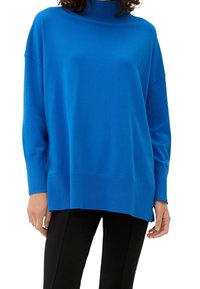 Femme portant un pull à col roulé bleu vif surdimensionné et un pantalon noir, les mains détendues le long du corps, sur un fond blanc.