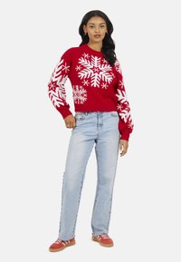 Pull en tricot rouge avec des motifs de flocons de neige blancs, assorti à un jean bleu clair à jambes droites et des baskets rouges à rayures blanches.