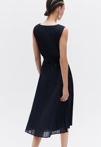 Abito midi plissettato blu navy con corpino aderente, design senza maniche e dettaglio in vita con fiocco. La texture liscia esalta la sua silhouette fluida.