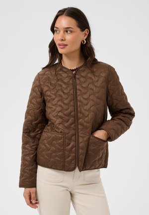 Femme portant une veste matelassée marron avec de grandes poches devant et un pantalon crème, debout avec une main dans une poche, regardant sur le côté.