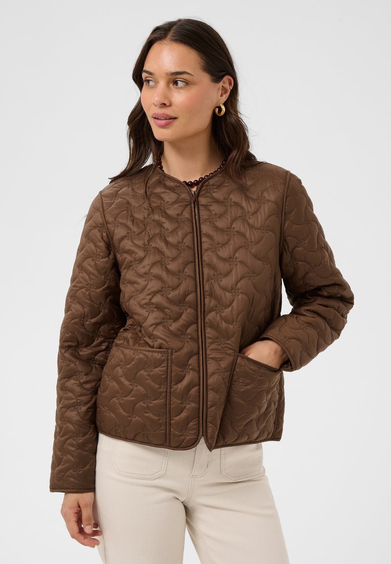 Femme portant une veste matelassée marron avec de grandes poches devant et un pantalon crème, debout avec une main dans une poche, regardant sur le côté.