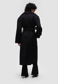 Černý vlněný trenčkot s páskem, širokými klopami a velkými knoflíky. Má rozparky vzadu a volný střih. Texturovaná tkanina s matným povrchem.