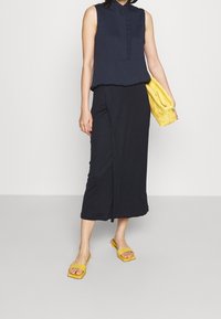 Námořnická bezrukávová blůza s vertikálními pruhy, s detailem knoflíku; černá midi sukně; žlutá pletená kabelka; žluté ploché sandály.