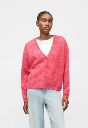 Vrouw draagt een zachtroze, pluizige cardigan over een wit shirt, gecombineerd met lichtblauwe spijkerbroek, staand tegen een effen lichte achtergrond.