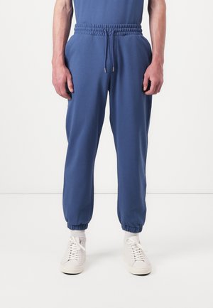 Blauwe sweatpants met elastische tailleband en manchetten, gemaakt van zachte stof, met een losse pasvorm en zijzakken, gecombineerd met witte sneakers.