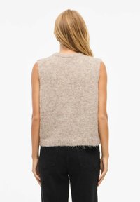 Sieviete ar gariem blondiem matiem, apģērbta bezapkaklīšu bēšā trikotāžas vestē un melnās biksēs, stāvot ar muguru pret baltu fonu.