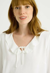 Blusa blanca con un collar volado y un lazo de cierre en el escote. Textura suave, tejido ligero y ajuste relajado alrededor de los hombros.