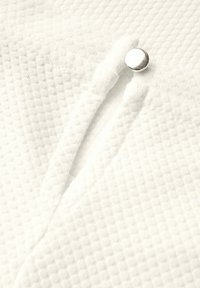 Tissu blanc texturé avec un motif en losanges, dotée d'un bouton en argent visible et de deux fentes verticales. Matériau doux et tissé.