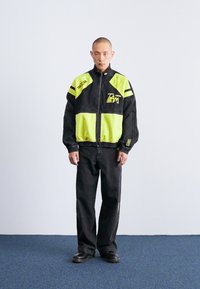 Casaco preto com detalhes e logótipos em amarelo-neon, combinado com calças pretas. O casaco tem fecho à frente e punhos elásticos.