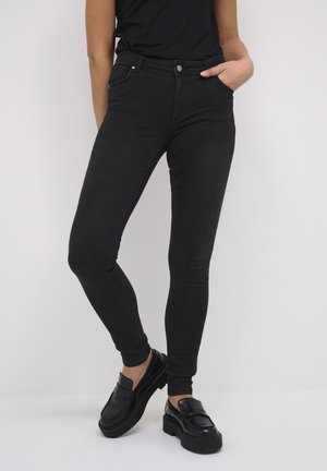 Personne portant un jean skinny noir et des mocassins à plateforme noirs, debout avec une main dans la poche sur un fond uni.