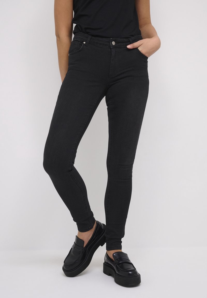 Personne portant un jean skinny noir et des mocassins à plateforme noirs, debout avec une main dans la poche sur un fond uni.