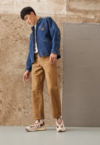 Jeansjacka med framfickor, khakifärgade rakbenade byxor och krämfärgade sneakers med röda detaljer. Texturerad väggbakgrund.