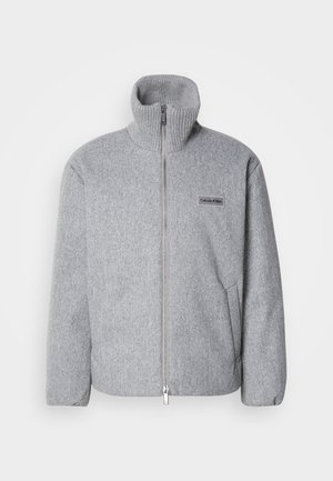Graue Zip-Jacke mit hohem Kragen, aus strukturierter Stoff gefertigt. Verfügt über Seitentaschen und ein Logo-Patch auf der Brust. Leichtes Design.