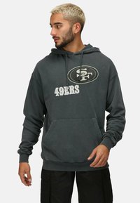 Sudadera de color gris oscuro con un bolsillo tipo canguro en la parte frontal, con un logo circular y el texto "49ERS" en blanco. Fabricada con un material suave.