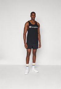 Top nero senza maniche con il logo "Champion" in bianco, abbinato a pantaloncini neri. Sneakers bianche e calzini alla caviglia a righe con accenti blu e gialli.