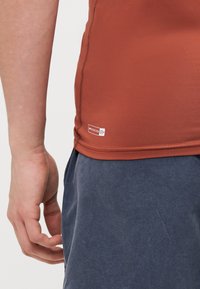 Kortermet, rustfarget treningsshirt laget av glattholdig stoff, med UV-beskyttelse-etikett og sydd kant, kombinert med marineblå shorts.