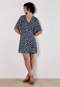 Marineblaues Kleid mit weißem Herzmuster, V-Ausschnitt, kurzen, ausgestellt geschnittenen Ärmeln und Knopfleiste; hat einen leichten Schlitz am Saum.