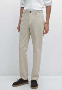 Pantaloni in cotone beige con un design a gamba dritta, tasche frontali e chiusura a un bottone, abbinati a scarpe in suede marrone scuro.