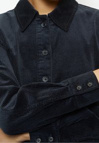 Giacca di velluto a coste nera con colletto a punta, bottoni frontali e maniche con bottoni, che mette in evidenza una superficie testurizzata e rigata.