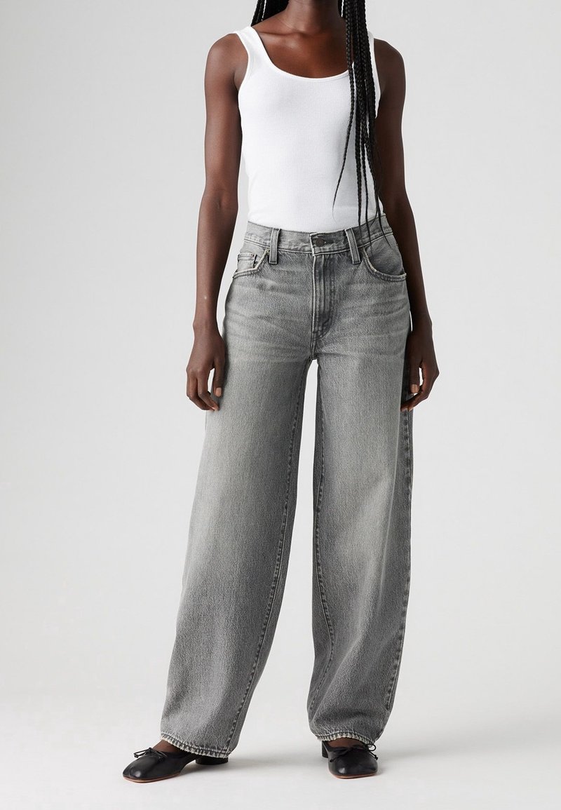 Personne portant un débardeur blanc, un jean gris taille haute à jambes larges et des chaussures plates noires, debout devant un fond uni.