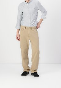 Beige Baumwoll-Chinos mit geradem Schnitt, kombiniert mit einem hellblau-weiß gestreiften Hemd und schwarzen Turnschuhen.