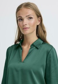 Camicia in satin verde con scollo a V e colletto, maniche lunghe e una texture liscia, indossata da un modello.