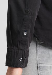 Maglietta nera a maniche lunghe con polsini bottonati; presenta un logo ricamato scuro e dettagli dei bottoni in contrasto. Tessuto di trama liscia.