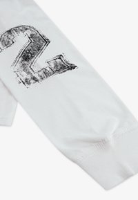 Valge puuvillane pikkade varrukatega särk, mille varrukal on must tekstuuriga number "2". Õmblused on nähtavad manseti juures.