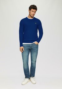 Blauer strukturierter Pullover mit einem weißen Rundhalsausschnitt, kombiniert mit hellblauen Jeans und weißen Sneakern. Verfügt über ein kleines Logo auf der Brust.