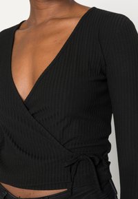 Schwarzes geripptes Wickeloberteil mit tiefem V-Ausschnitt und langen Ärmeln; verfügt über ein seitliches Bindedetail an der Taille zur Anpassung.