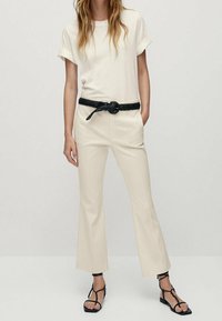 T-shirt crema con maniche arrotolate, abbinata a pantaloni beige a vita alta con tasche laterali e una cintura nera intrecciata. Sandali neri.