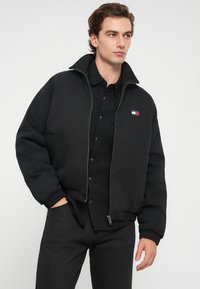 Black bomber jacket with a high collar, zip closure, ribbed cuffs, and embroidered logo patch. Worn over a black shirt and pants.