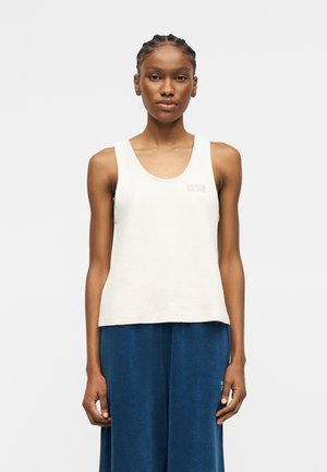 Γυναίκα με πλεξούδες που φοράει λευκό αμάνικο τοπ και άνετο μπλε παντελόνι, στέκεται μπροστά σε απλό φωτεινό φόντο.