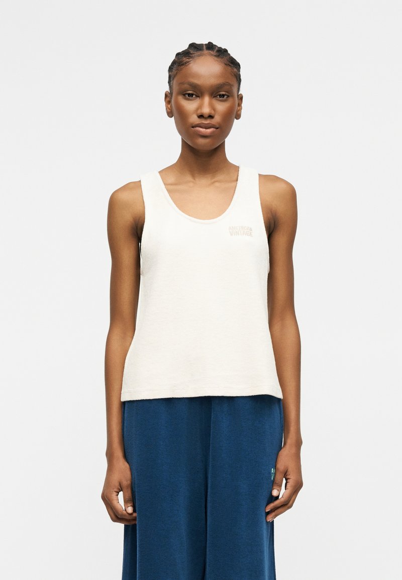 Γυναίκα με πλεξούδες που φοράει λευκό αμάνικο τοπ και άνετο μπλε παντελόνι, στέκεται μπροστά σε απλό φωτεινό φόντο.