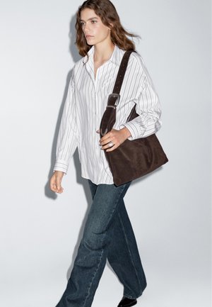 Camicia bianca a righe con bottone, jeans scuri a zampa d'elefante e una borsa a spalla in suede marrone con un manico caratterizzato da una fibbia argento.