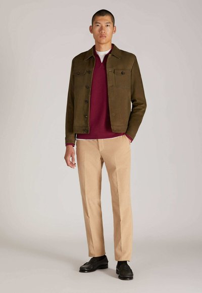 Giacca verde oliva con quattro tasche frontali, indossata sopra un maglione bordeaux e una maglietta crema. Pantaloni beige e scarpe nere completano l'outfit.