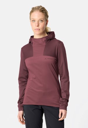 Frau mit blondem zurückgebundenem Haar, die ein weinrotes Langarm-Kapuzenoberteil und schwarze Hose trägt, steht vor einem schlichten hellen Hintergrund.