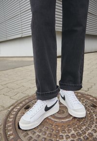 Bijele Nike tenisice s crnim Swoosh-om, sivosmeđi vrh i bijele vezice u kombinaciji s tamnim trapericama s podvrnutim rubovima na uzorkovanoj površini.